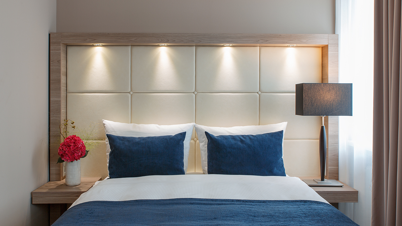 Helles Hotelzimmer mit Bett und blauer Tagesdecke sowie blauen Kissen und Nachtischlampe