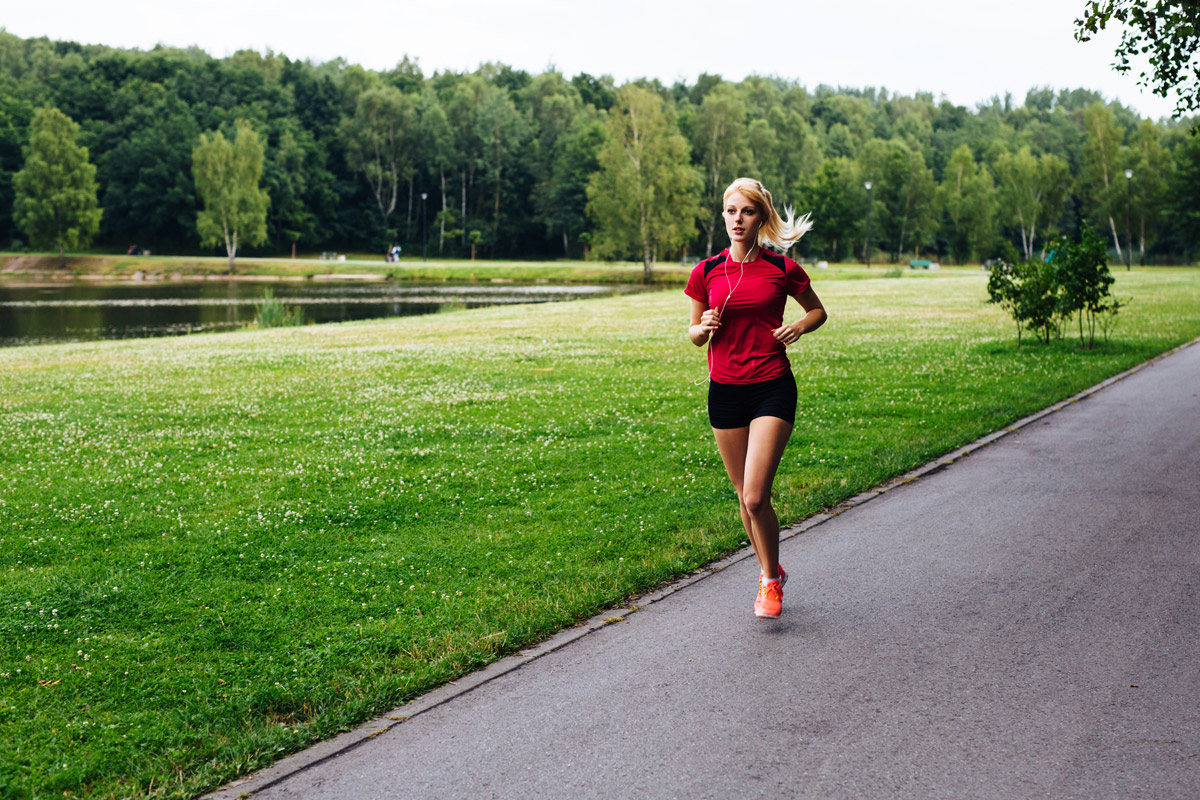 Junge, blonde Frau in Sportkleidung vor einer gemähten Wiese joggt und dabei Musik hört