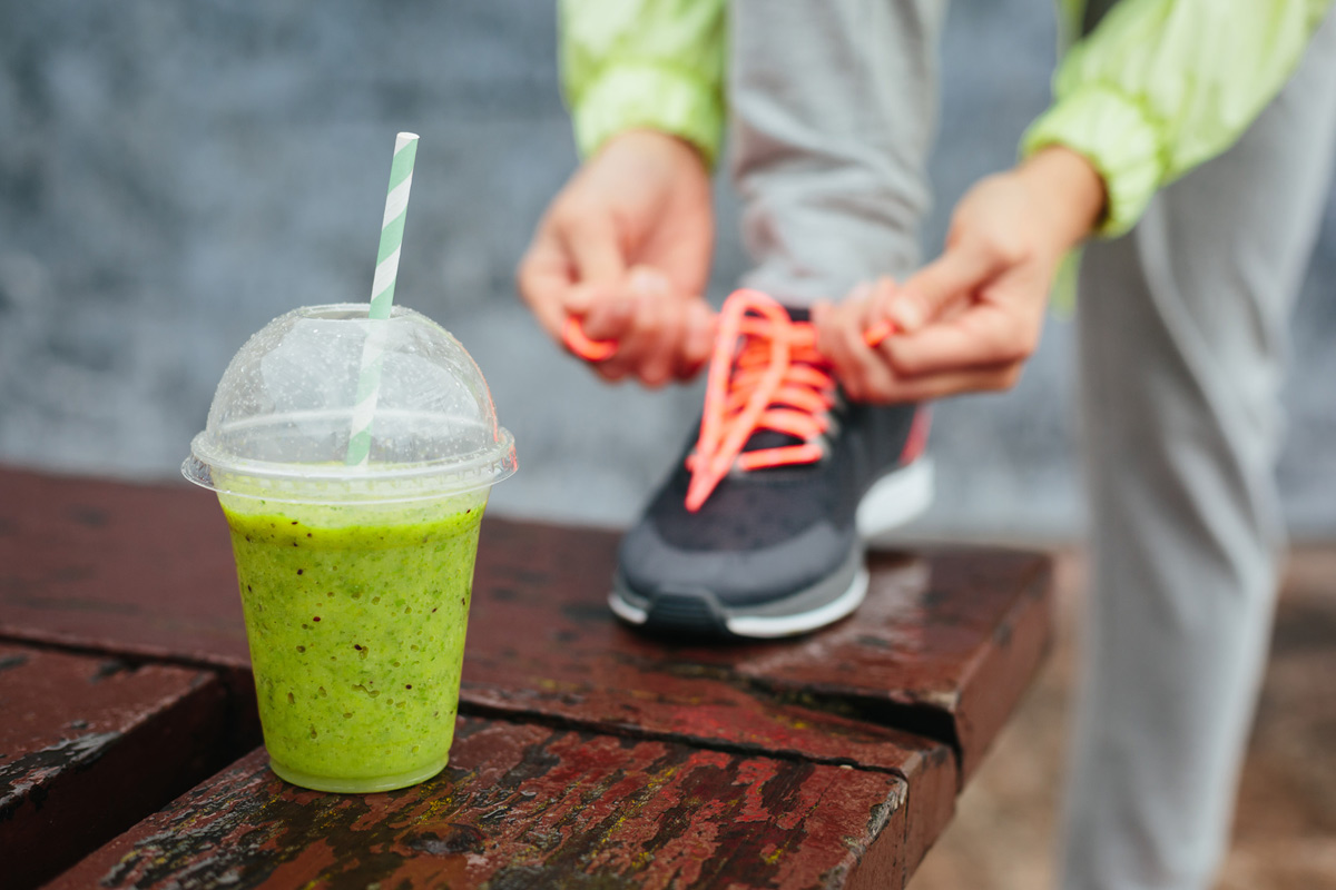 Grüner Smoothie hinter dem sich ein Sportler in Tiefenschärfe den Turnschuh bindet