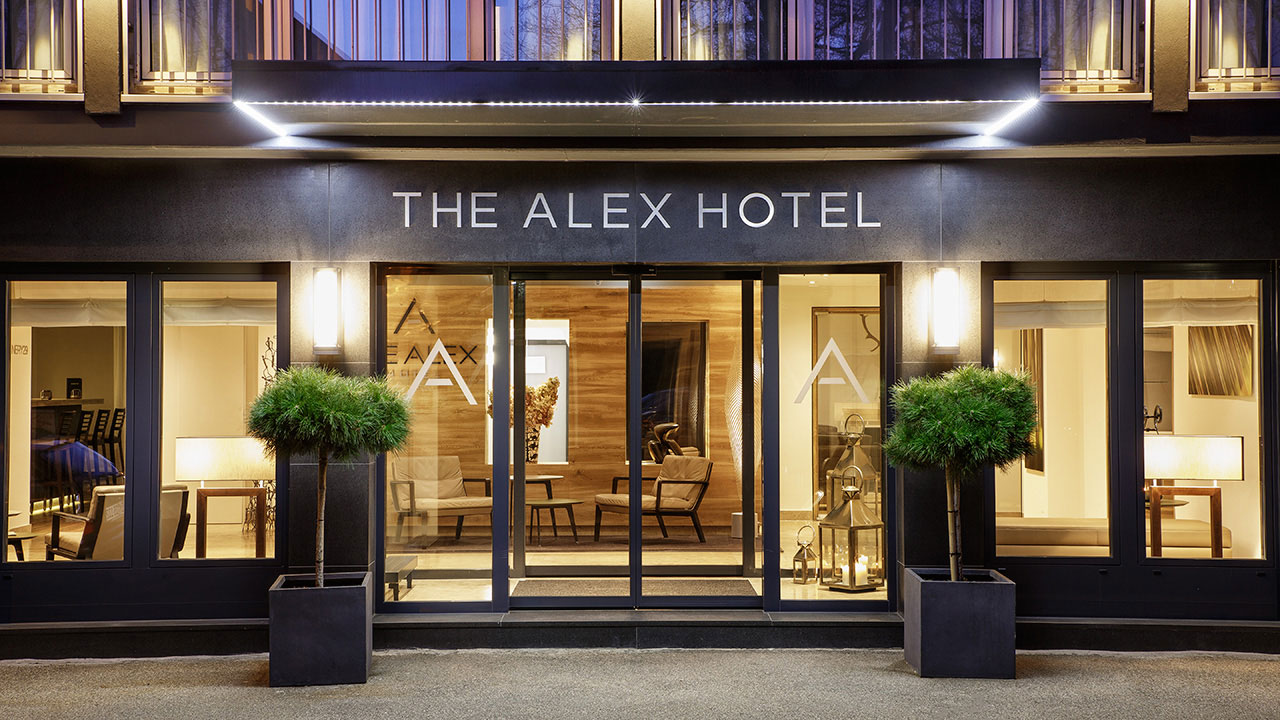 Eingangsbereich von The Alex Hotel von außen, Glasfront mit zwei kleinen Bäumen neben der Eingangstür