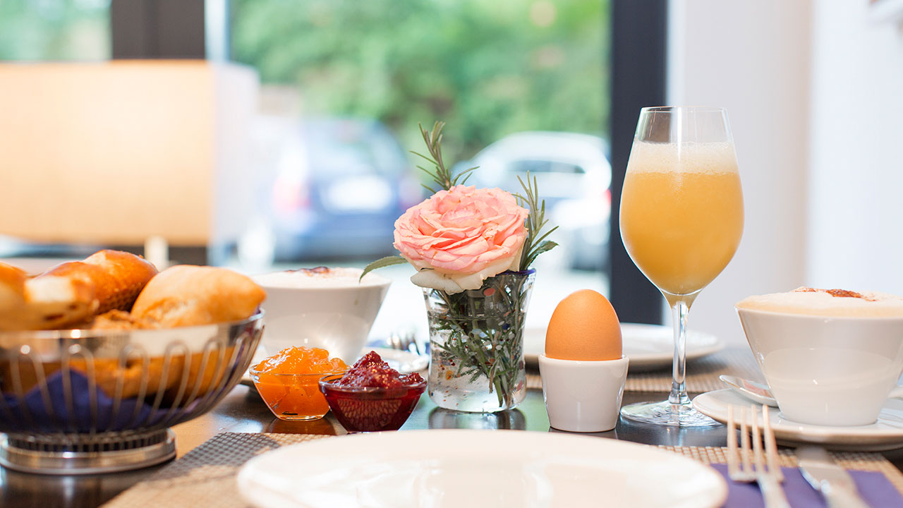 Reichlich gedeckte und mit Blumen dekorierter Frühstückstisch mit Orangesaft, Marmelade, Frühstücksei, Kaffee und Cappuccino