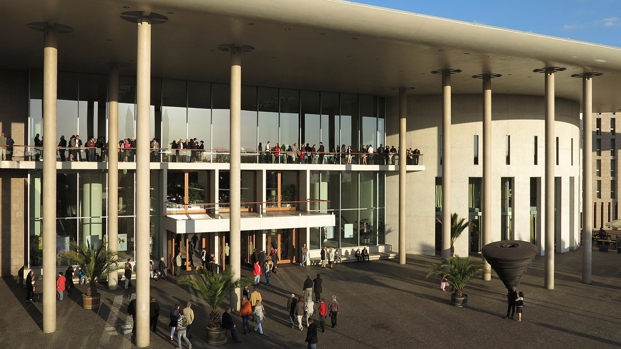 Überdachter Eingangsbereich mit Balkon darüber und mehreren dünnen Säulen sowie vielen Besuchern